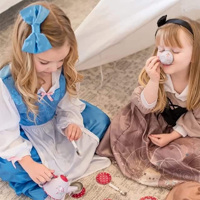 Meisje draagt een blauwe Belle jurk kind met witte schort, lange mouwen en grote blauwe haarstrik tijdens een theekransje.