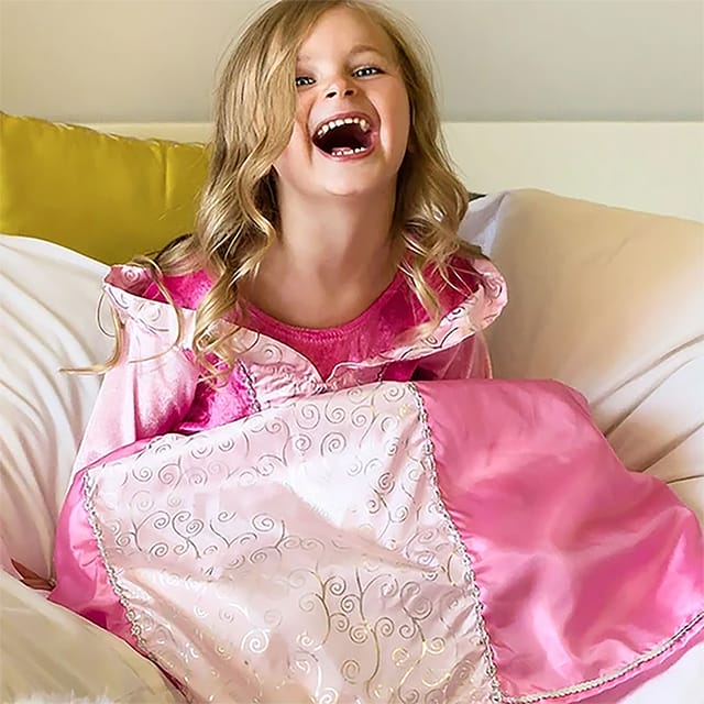 Lachend meisje draagt een roze Doornroosje jurk met witte sierkrullen, zittend op een bed