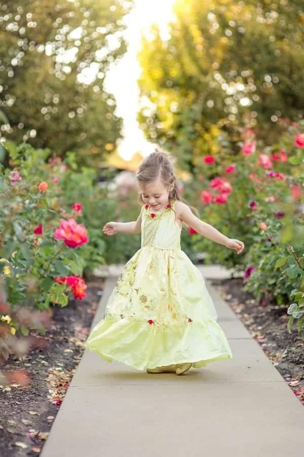 Meisje draagt een gele Belle prinsessenjurk met rozen terwijl ze door een bloeiende tuin loopt