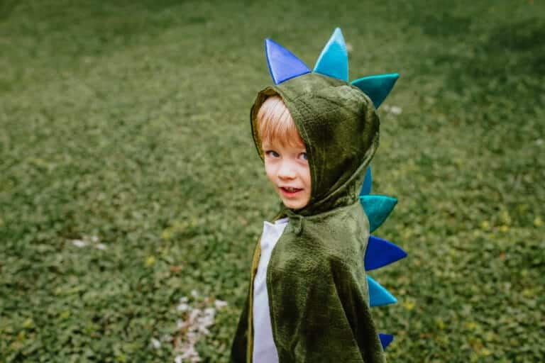 Jongen draagt een groene drakencape met capuchon en blauwe stekels, buiten op gras van opzij gezien