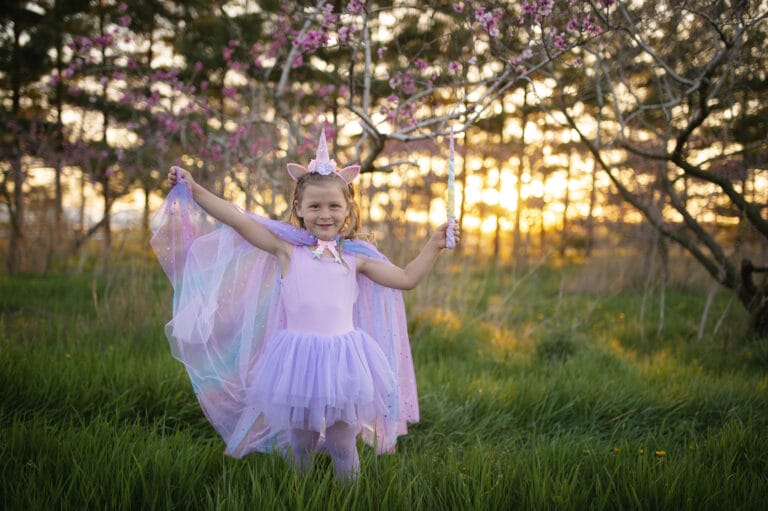 Meisje draagt een paarse unicorn cape met tule en eenhoornhaarband in een bloeiende tuin bij zonsondergang.
