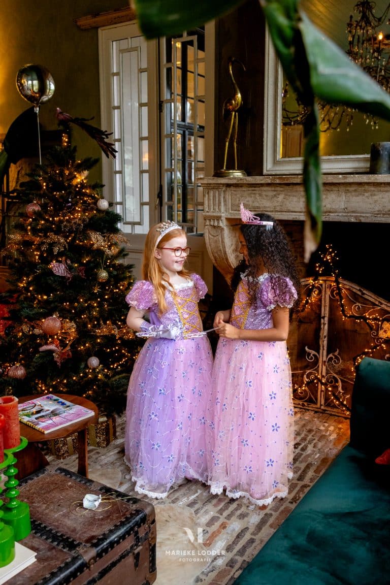 Twee kinderen dragen de paarse en roze Rapunzel prinsessenjurken met bloemenprint en glitterdetails naast een kerstboom.