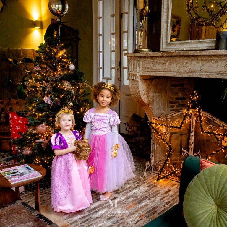 Twee meisjes dragen roze Rapunzel-jurken met Rapunzel accessoires en kroontjes naast een verlichte kerstboom.