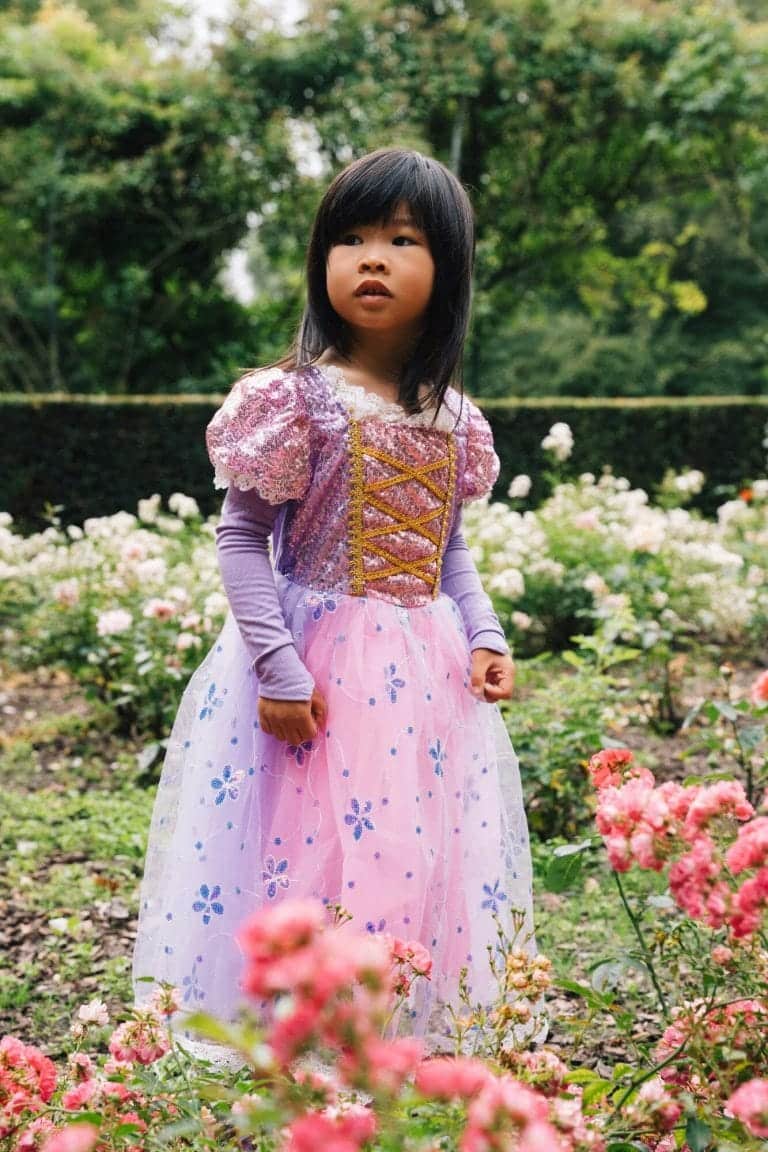 Meisje draagt een paarse en roze Rapunzel verkleedjurk met gouden veterdetails in een bloeiende tuin