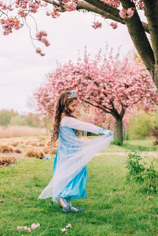 Meisje draagt een blauwe prinsessenjurk uit het Frozen pakket met witte, doorschijnende mouwen in een bloeiende tuin.