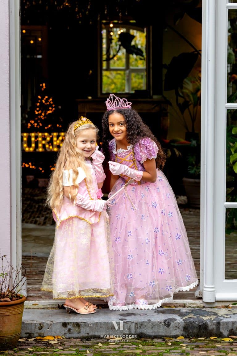 Twee meisjes dragen roze prinsessenjurken van Doornroosje en Rapunzel met tiara’s, handschoenen en toverstaf bij een deur
