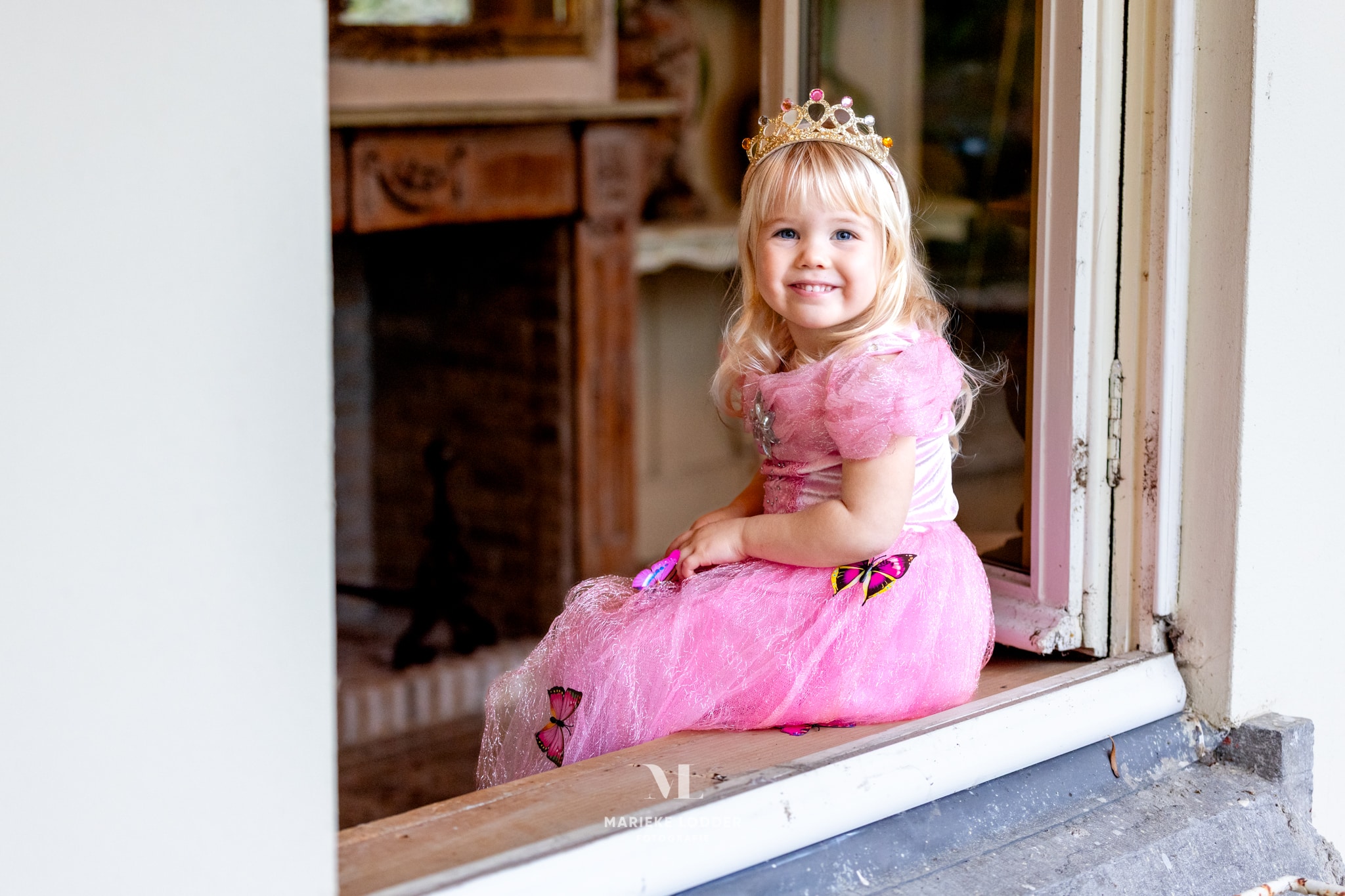 Zittend in een deuropening lacht een kind in een roze Doornroosje jurk met vlinderversiering en gouden tiara.