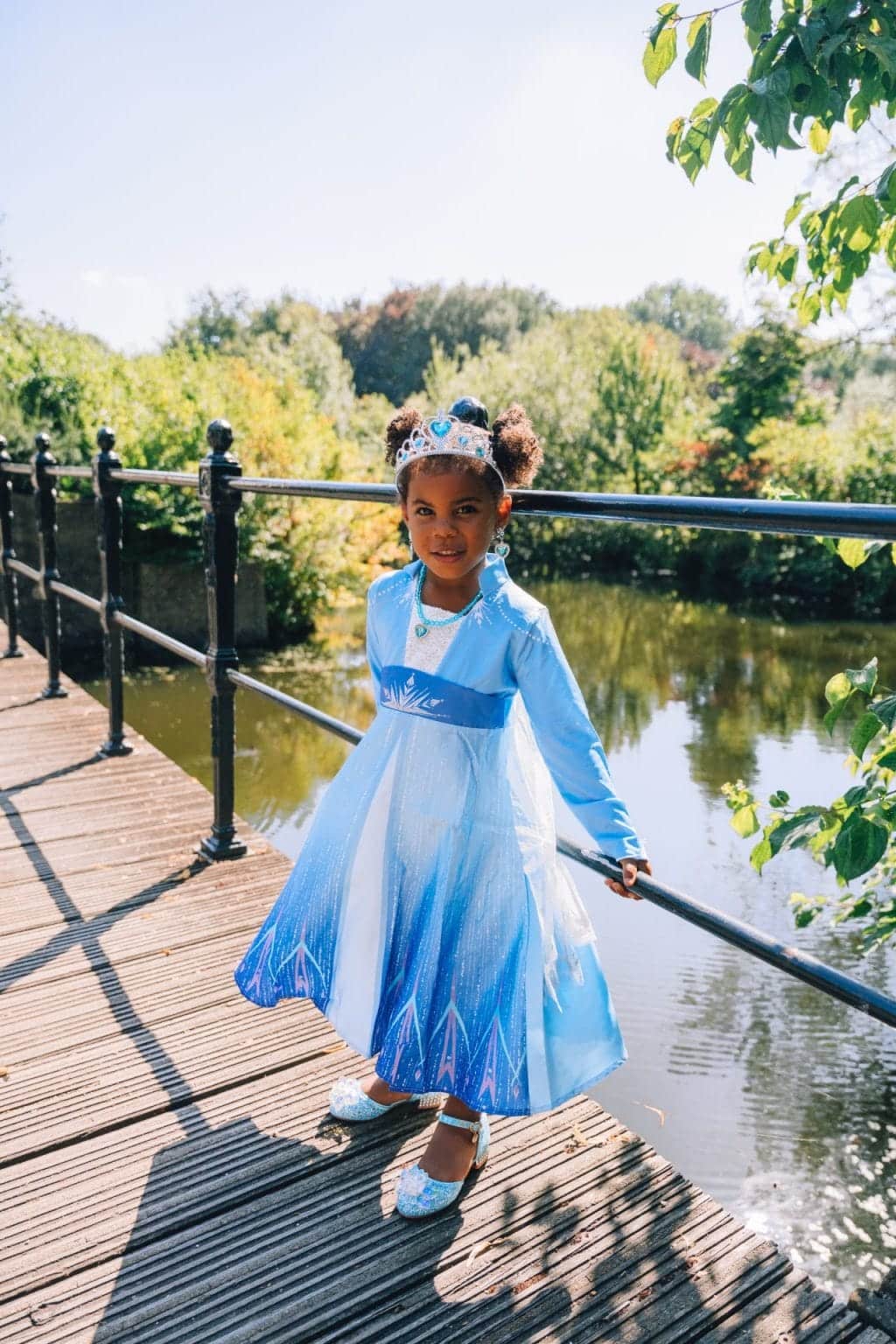 Meisje draagt een blauwe jurk frozen elsa met glanzende rok en kroon op een houten brug bij het water.