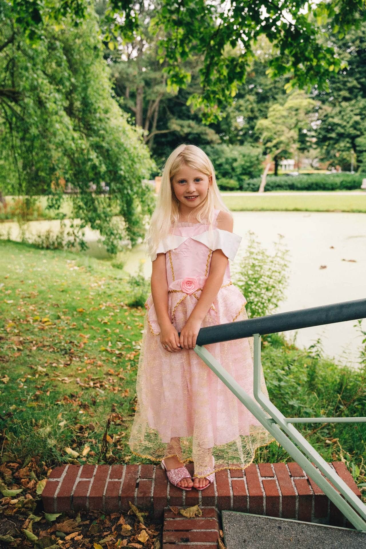 Meisje draagt een roze Doornroosje prinsessenjurk met roos en gouden details bij een brug in het park