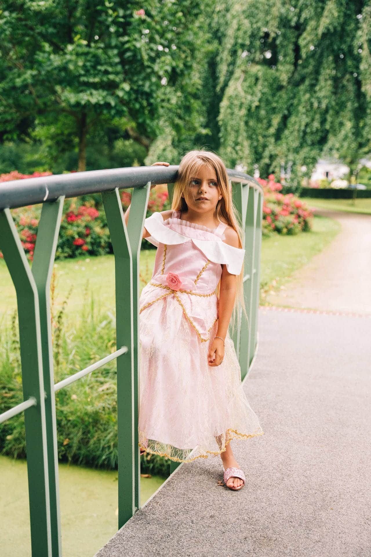 Op een brug in een bloeiende tuin staat een meisje in de roze Doornroosje jurk, van voren zichtbaar met roos op de taille.