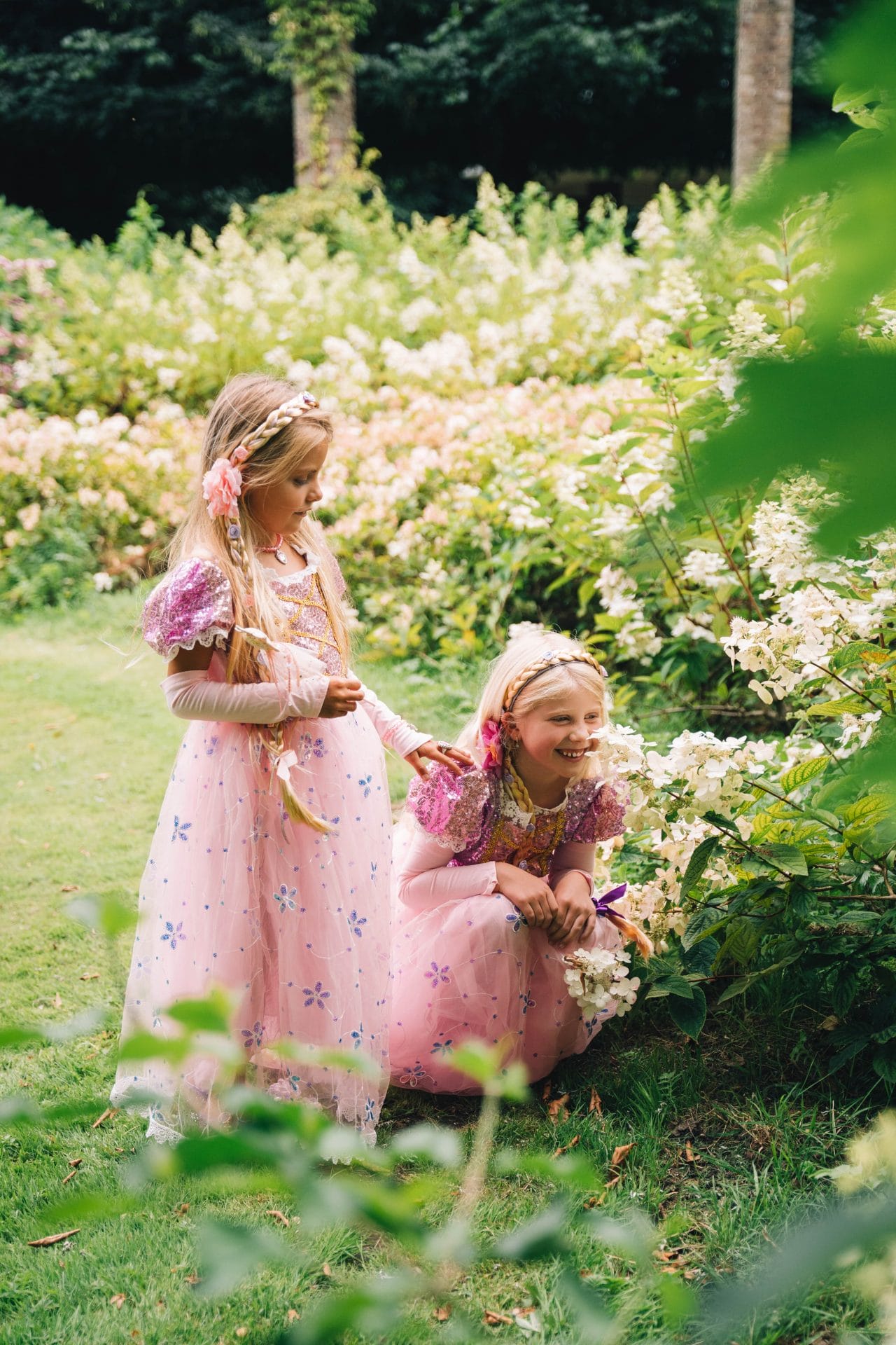 Twee meisjes dragen roze Rapunzel jurken met bloemenprint en paarse glittermouwen in een bloeiende tuin.