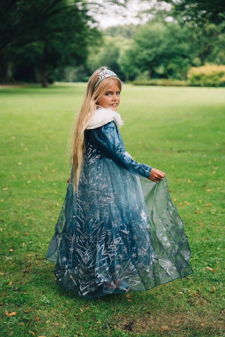 Meisje draagt een blauwe Frozen Elsa-prinsessenjurk met tiara, witte bontkraag en zilveren sneeuwvlokken in een park.