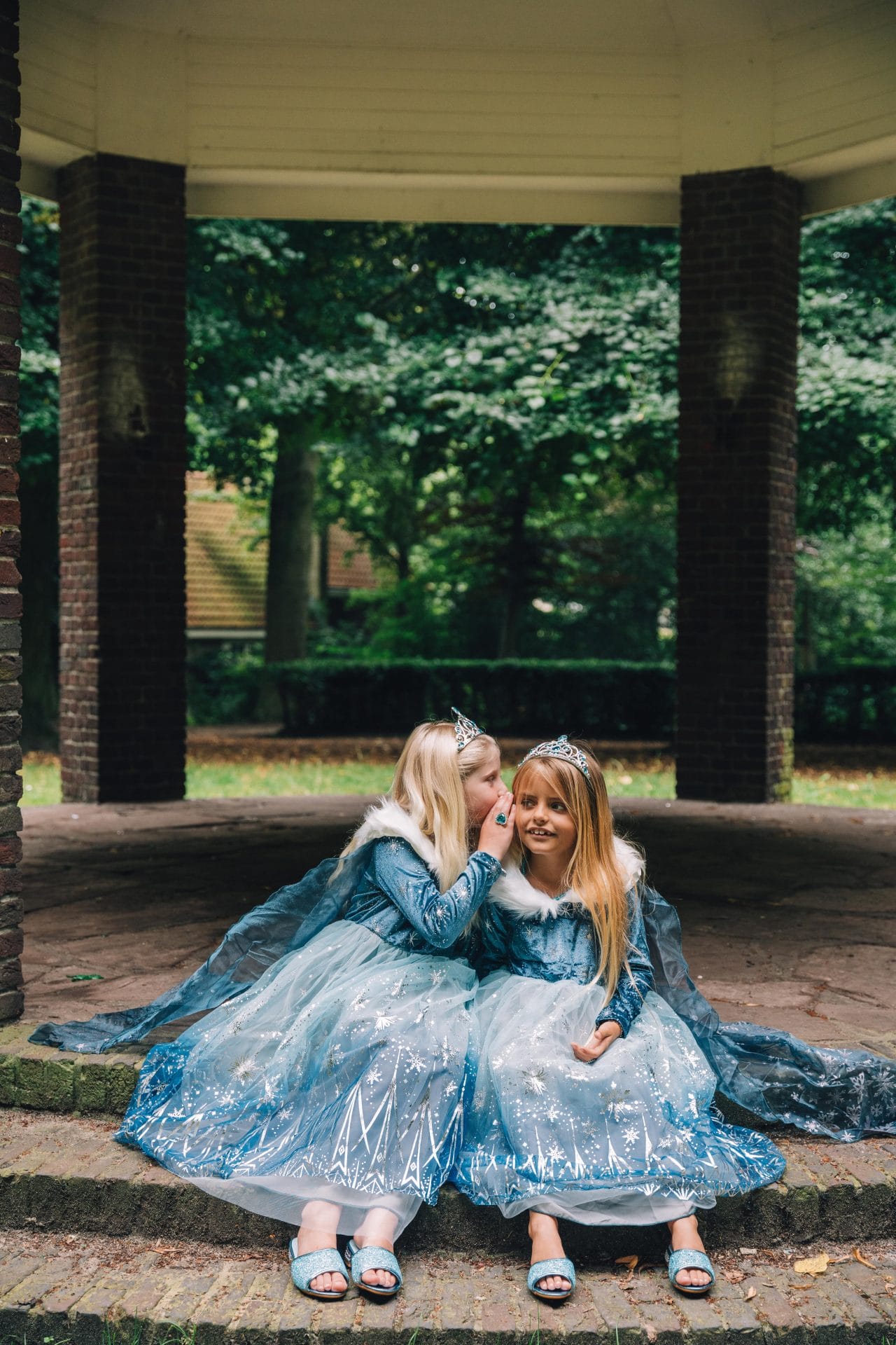 Twee meisjes dragen een blauwe frozen prinsessenjurk met tiara en glinsterende rok, terwijl één iets in het oor fluistert.