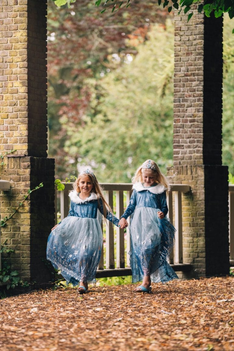 Twee meisjes dragen een blauwe elsa jurk met tiara, witte bontkraag en glinsterende rok, terwijl ze hand in hand lopen.