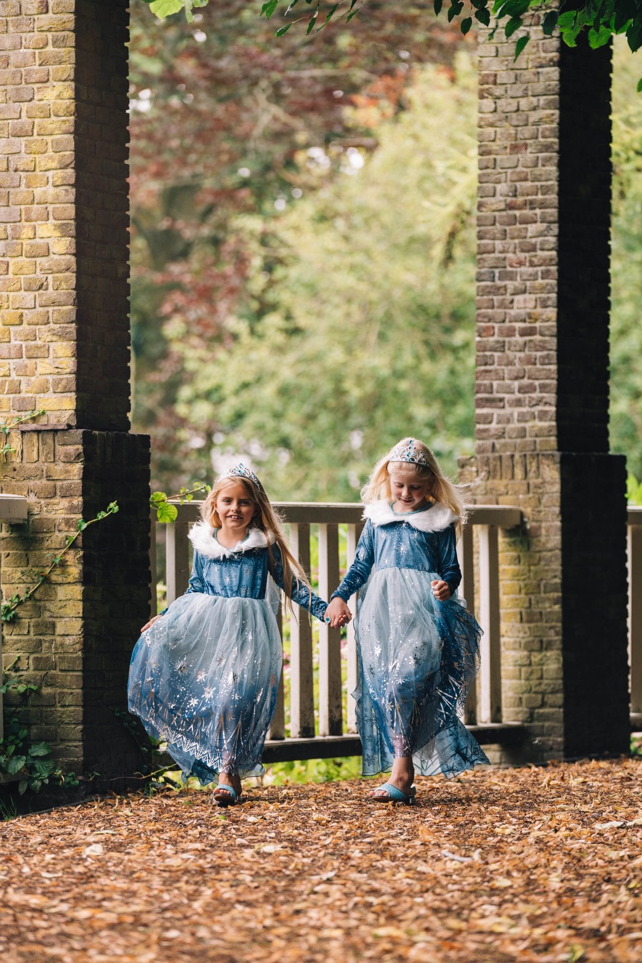 Twee meisjes dragen een blauwe elsa jurk met tiara, witte bontkraag en glinsterende rok, terwijl ze hand in hand lopen.