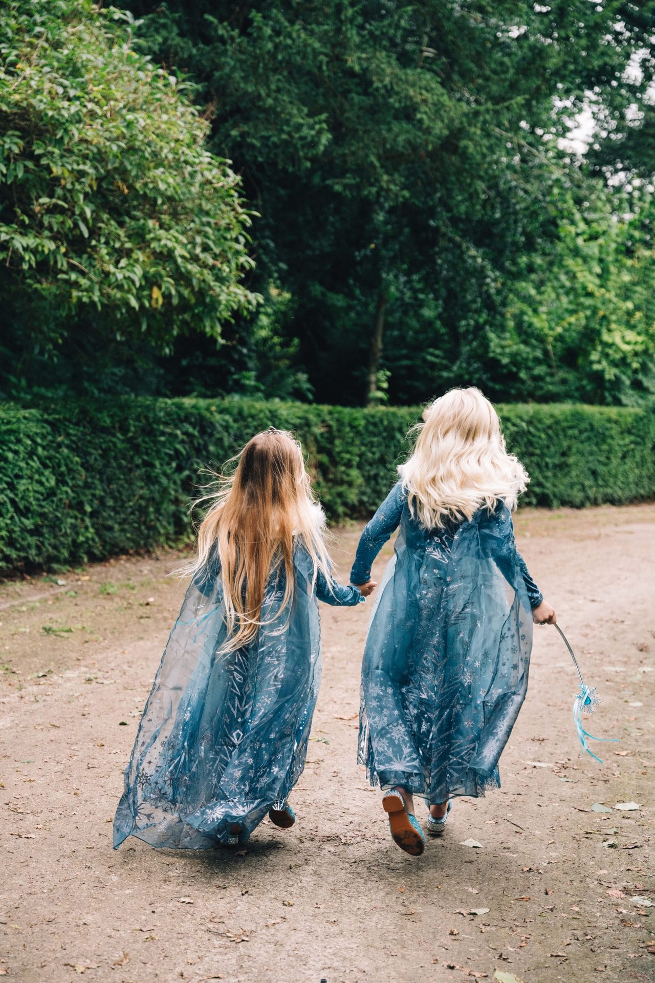 Twee meisjes dragen elk een blauwe luxe prinsessenjurk van Lunja en lopen hand in hand over een bospad.