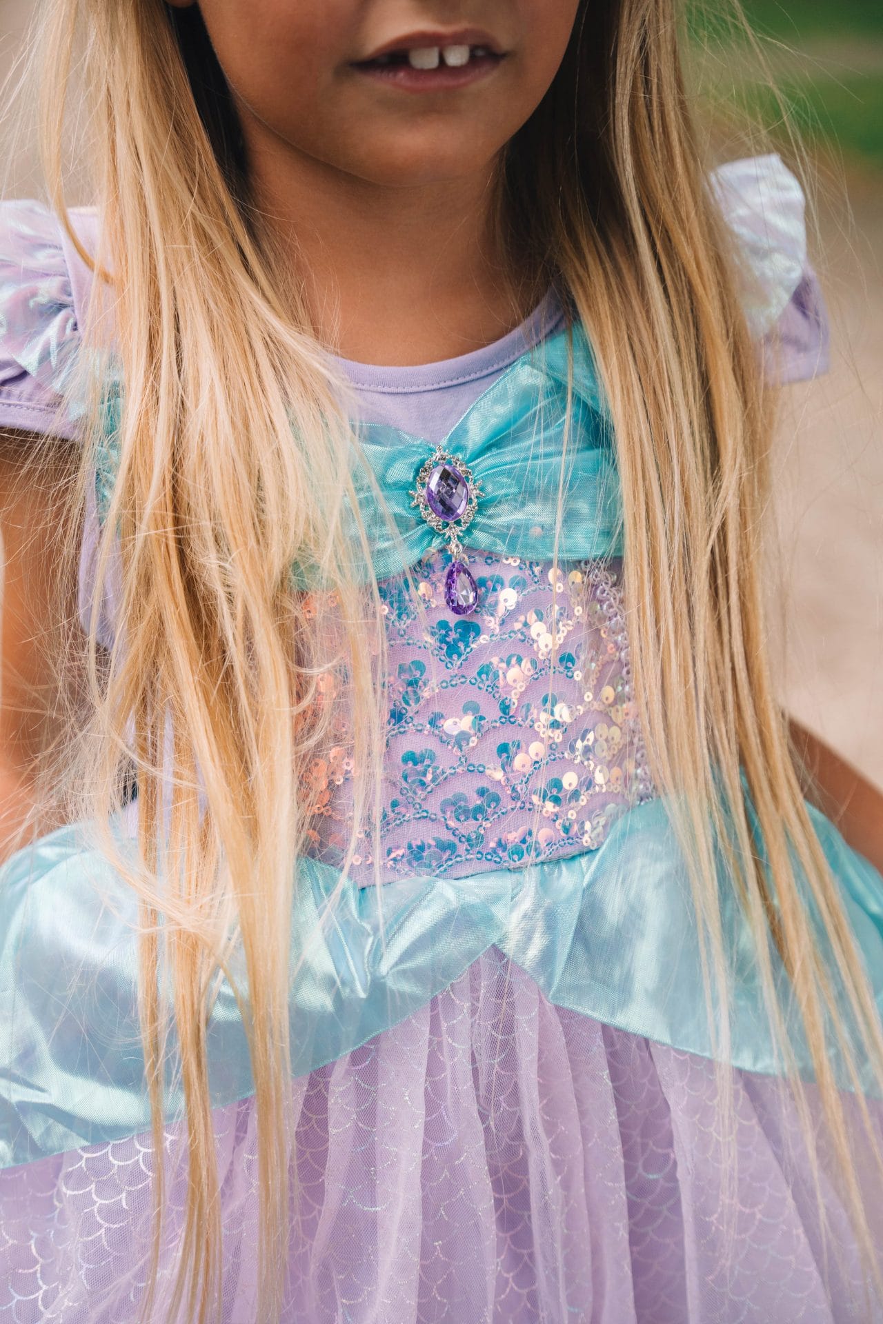 Close-up van een kind dat een turquoise en paarse jurk ariel draagt, met glanzend lijfje, sierstenen en schubbenpatroon.
