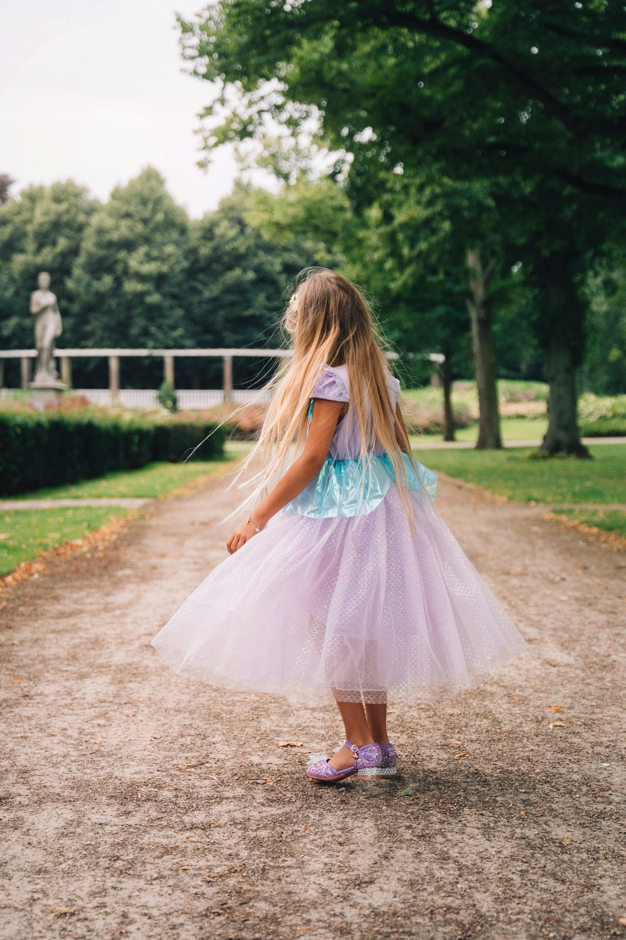 Achteraanzicht van een meisje in een paarse zeemeerminjurk met glinsterende schoenen op een pad in het park.