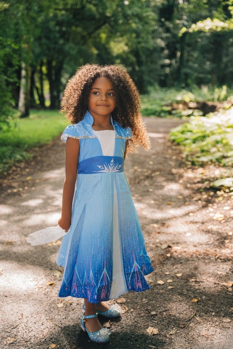 Meisje draagt een blauwe Elsa jurk met glinsterende rok en zilverkleurige versieringen op een bospad.
