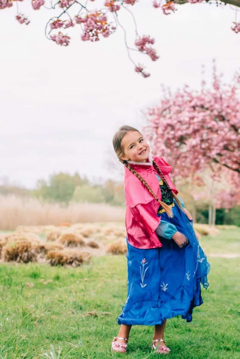 Meisje draagt een blauwe Anna jurk met roze cape en lange vlechten in een bloeiende tuin.