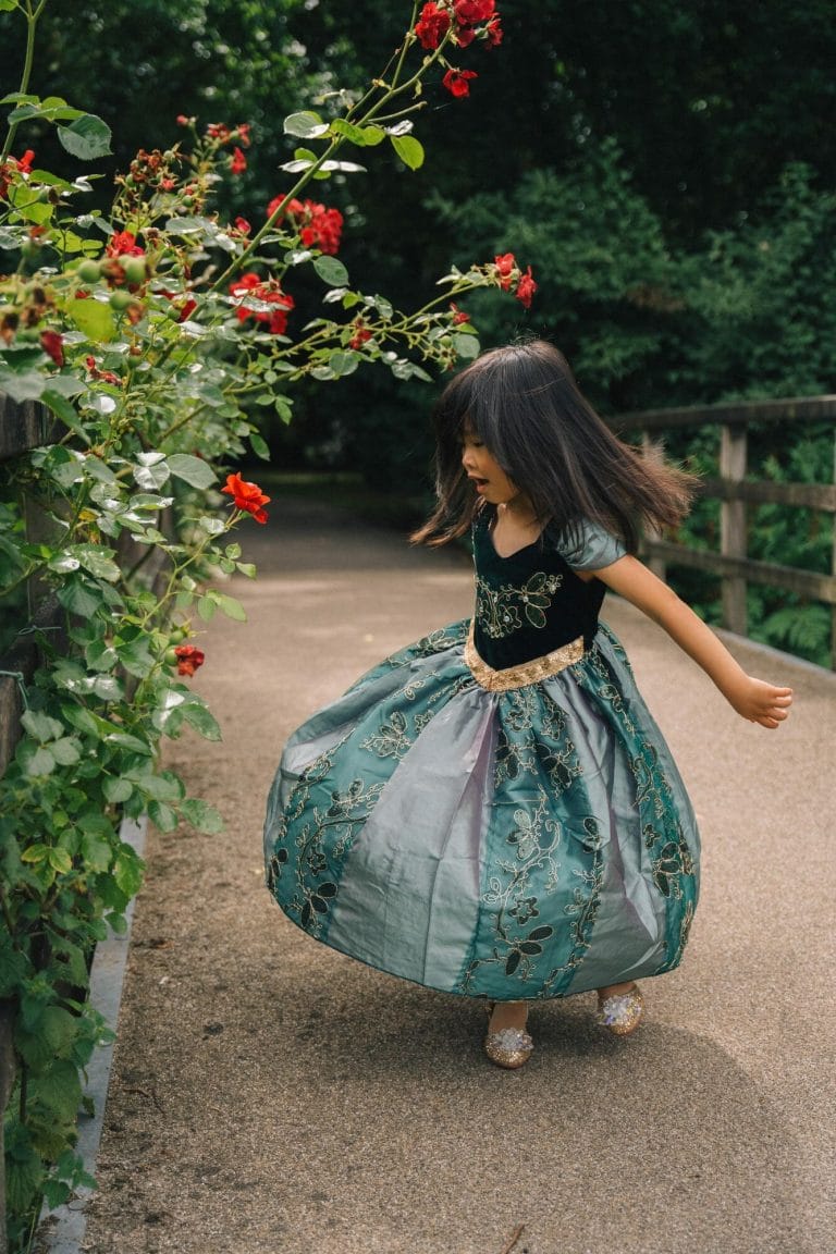 Meisje draagt een groene anna jurk met gouden bloemenborduursel en glanzende rok op een tuinpad.