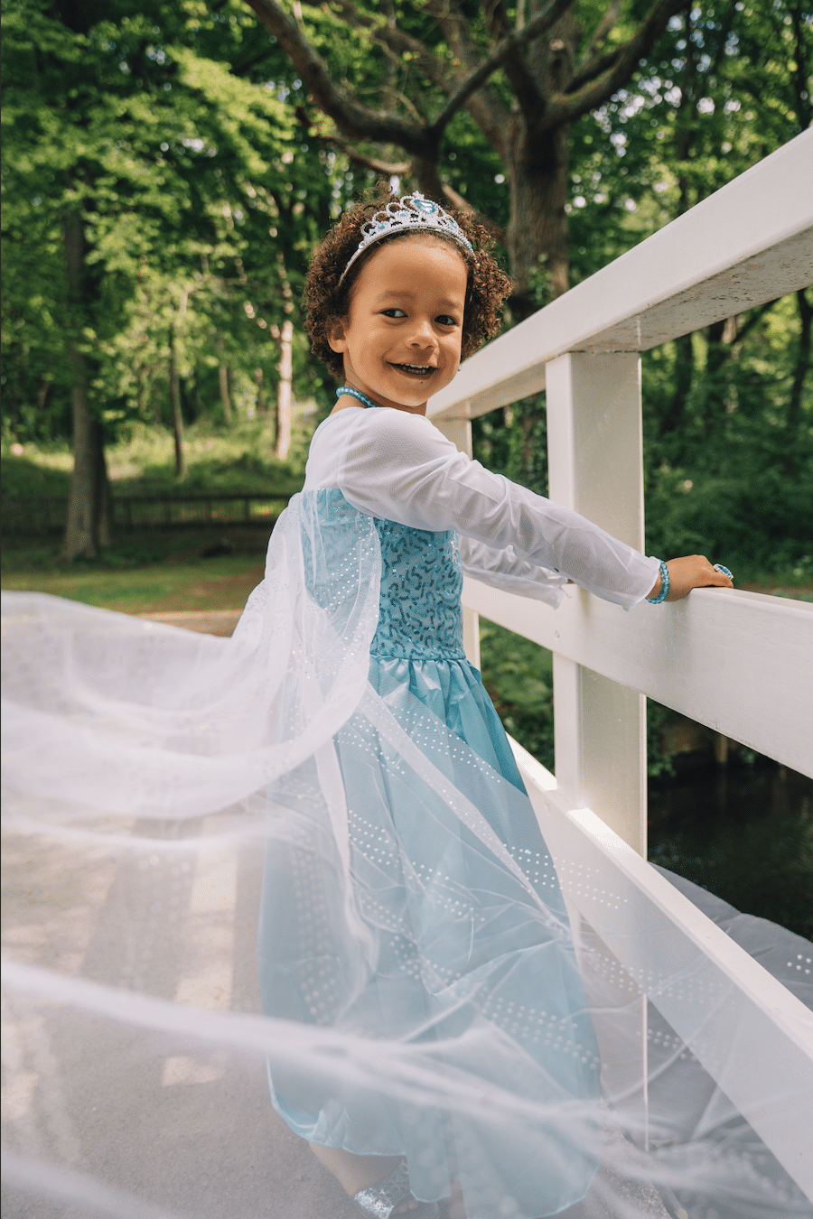 Meisje draagt een blauwe Elsa-prinsessenjurk met witte mouwen en tiara op een witte brug in het bos.