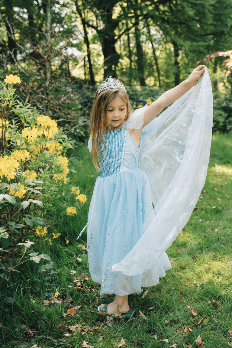 Meisje in een blauwe Elsa prinsessenjurk met tiara houdt de lange witte, doorschijnende cape omhoog in een tuin.