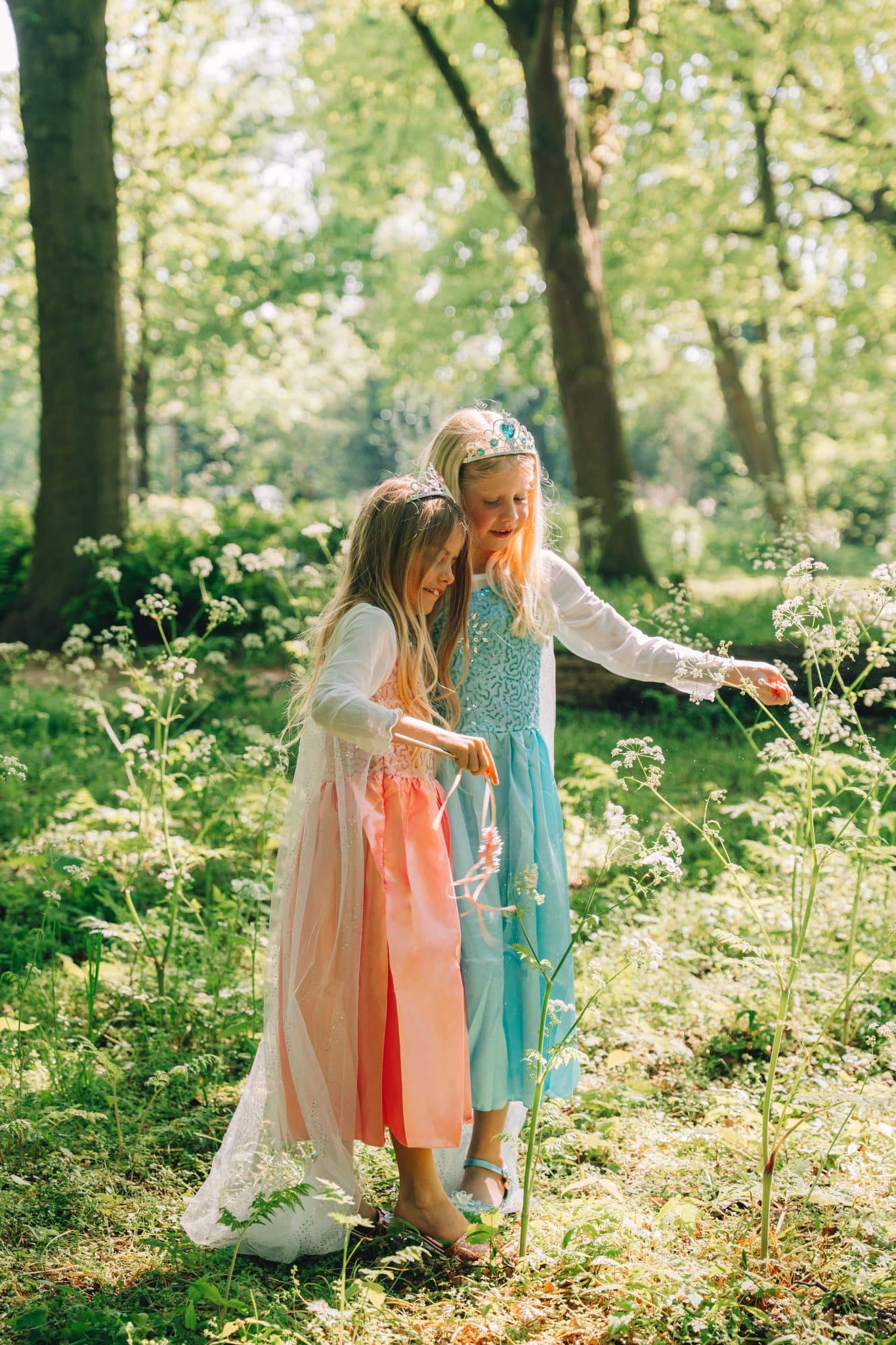 Twee meisjes dragen een blauwe prinsessenjurk en roze Doornroosje-jurk met tiara’s in een zonnig bos