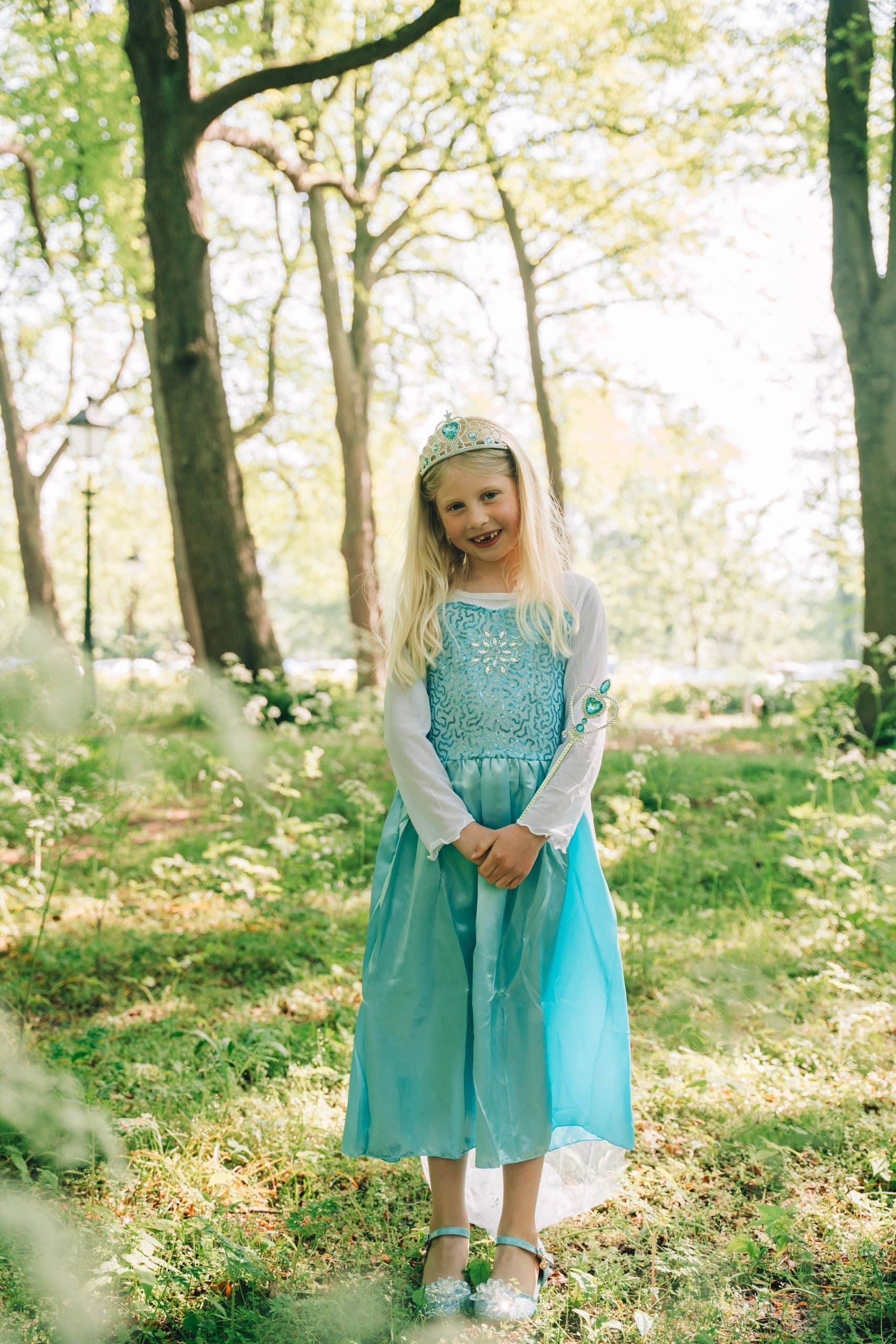 Meisje draagt een blauwe Frozen Elsa prinsessenjurk met tiara, witte mouwen en toverstaf in een zonnig bos.