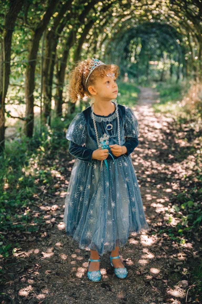 Op een bospaadje staat een kind in een blauwe Elsa prinsessenjurk, volledig van voren zichtbaar met tiara en toverstaf.