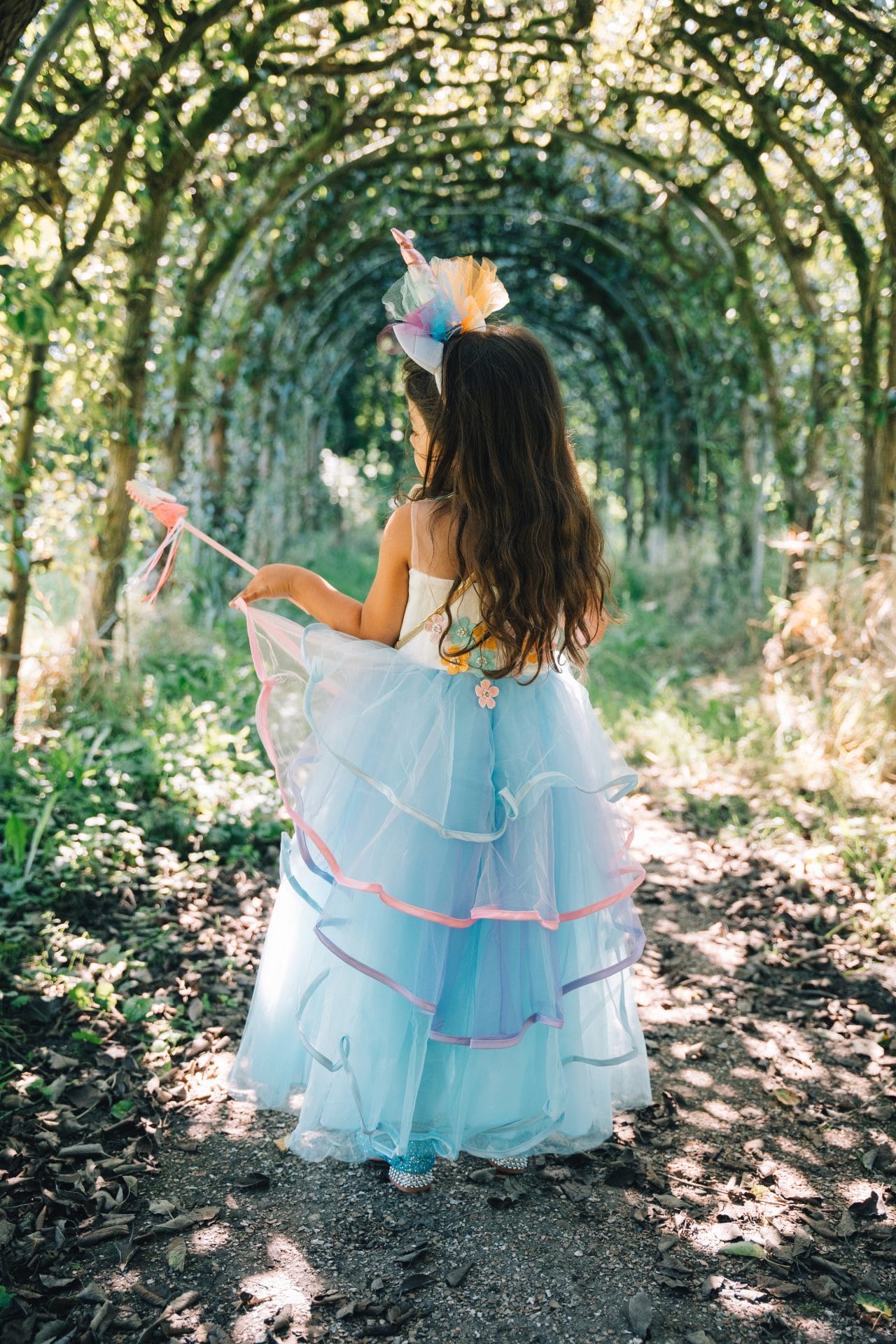Van achteren gezien draagt een meisje een blauwe unicorn jurk met tule rok en eenhoornhaarband in het bos.