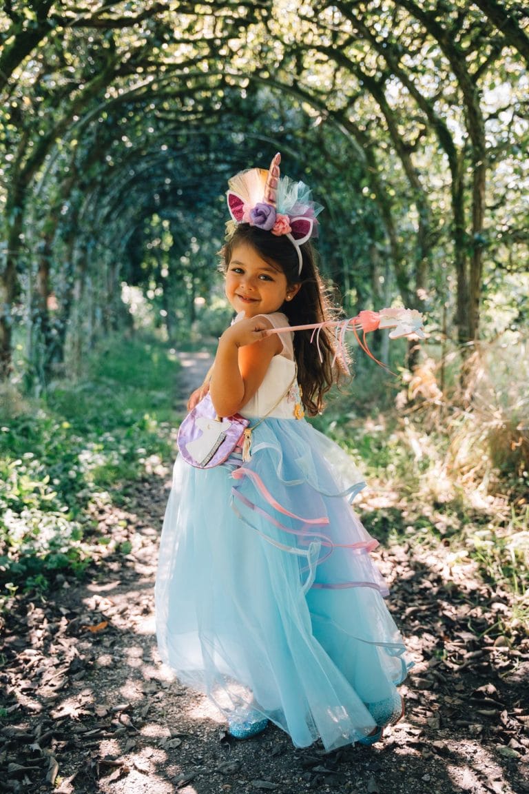 Meisje draagt een blauwe unicorn jurk met lange tule rok, eenhoornhaarband en roze toverstaf in het bos.