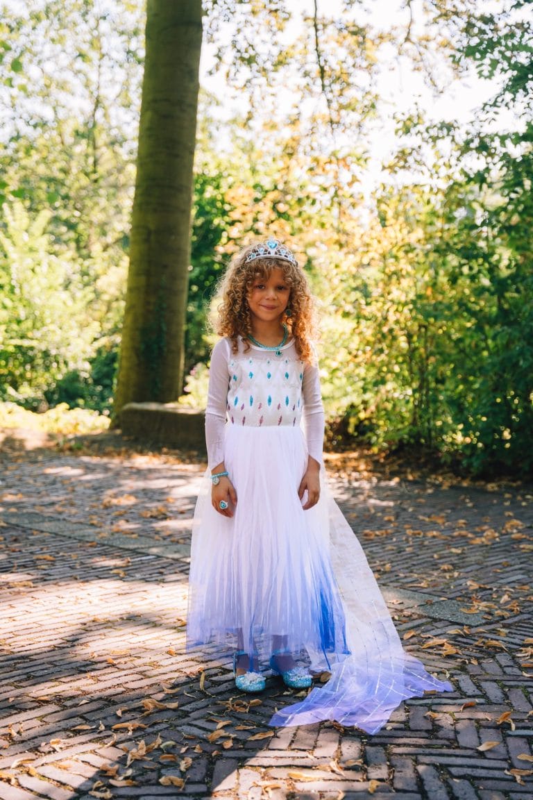 Meisje draagt een witte Elsa jurk met lila rok en lange doorschijnende sleep, kroon op het hoofd in een bos