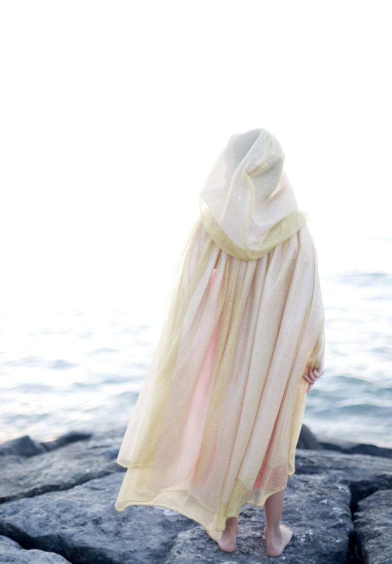 Kind van achteren in een roze en gouden prinsessen cape met capuchon op rotsen aan zee
