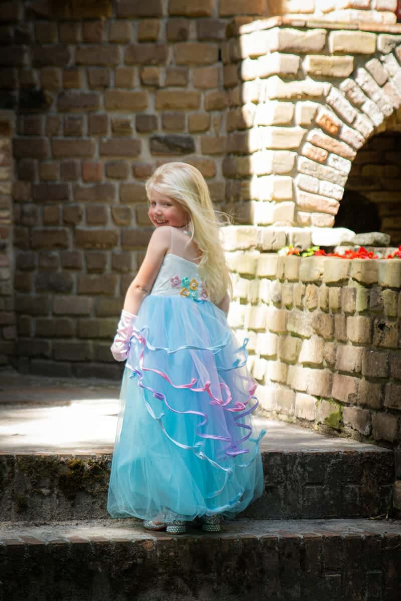Meisje draagt een blauwe unicorn jurk met lange tule rok, bloemen op de rug en roze en paarse linten bij een stenen muur.