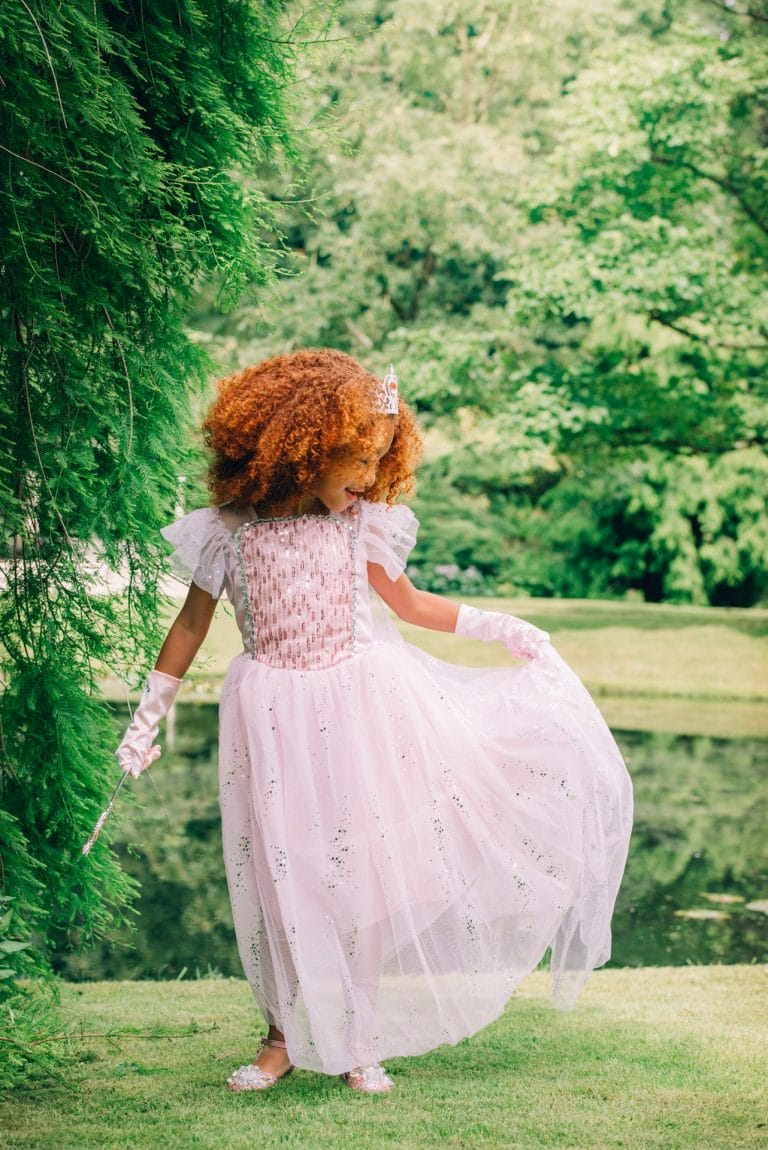 Een meisje draagt een roze Elsa prinsessenjurk van Het Betere Merk met tiara, handschoenen en toverstaf in een groene tuin.