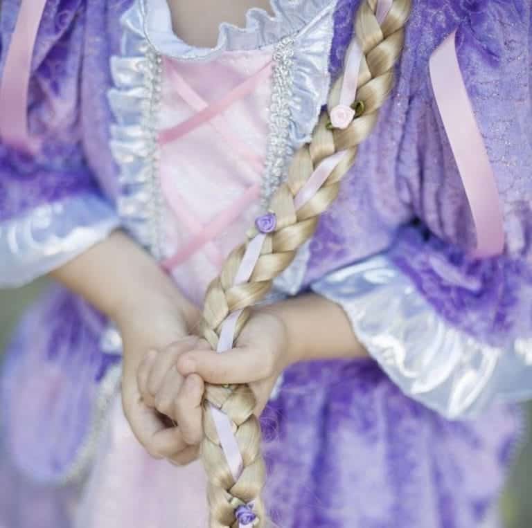 Kind in paarse jurk houdt de blonde Rapunzel haarvlecht van Great Pretenders vast, versierd met lila linten en bloemetjes.