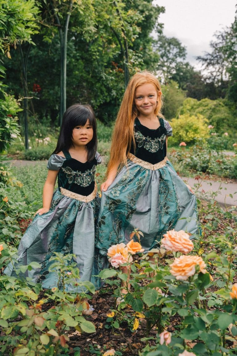 Twee meisjes dragen een Anna jurk met donker groen lijfje, geborduurde versiering en wijde blauwgroene rok in een rozentuin.