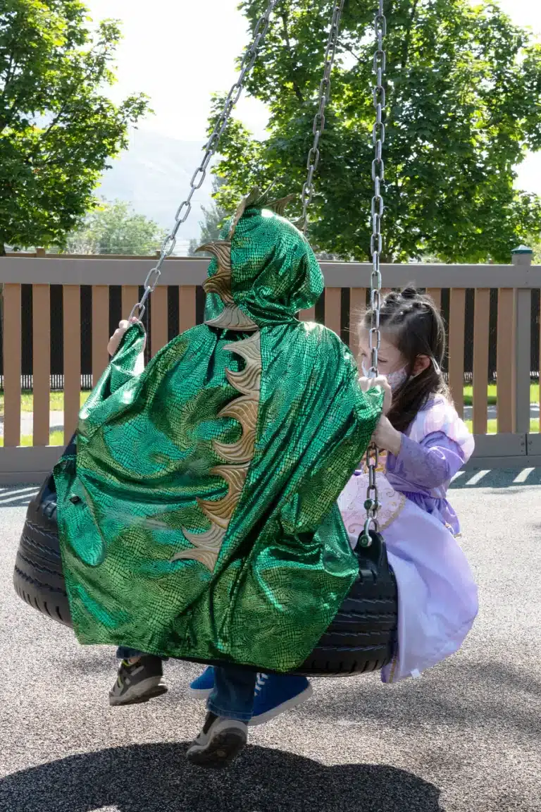 Een kind draagt een groen drakenpak op een schommel, gezien van achteren met goudkleurige stekels op de rug.