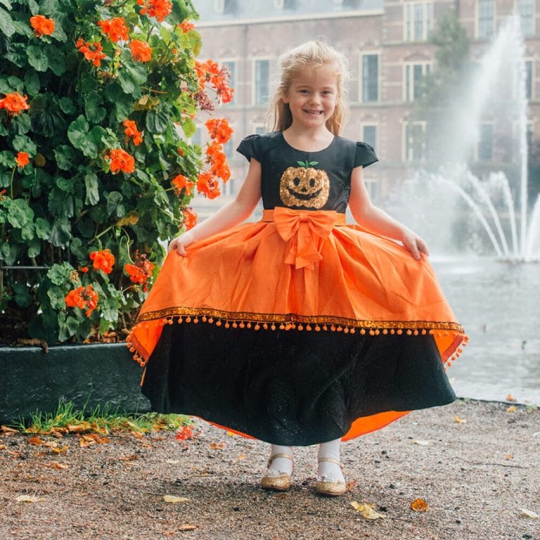 Meisje draagt een oranje-zwart halloween kostuum kind met pompoenprint, strik en wijde rok bij een fontein.