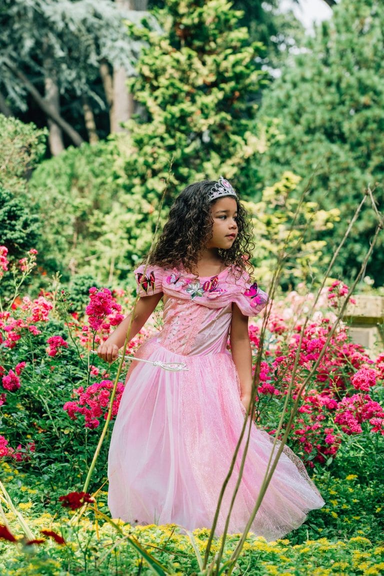 Meisje draagt een roze prinsessenjurk Doornroosje van Het Betere Merk met tiara en toverstaf in een bloementuin.