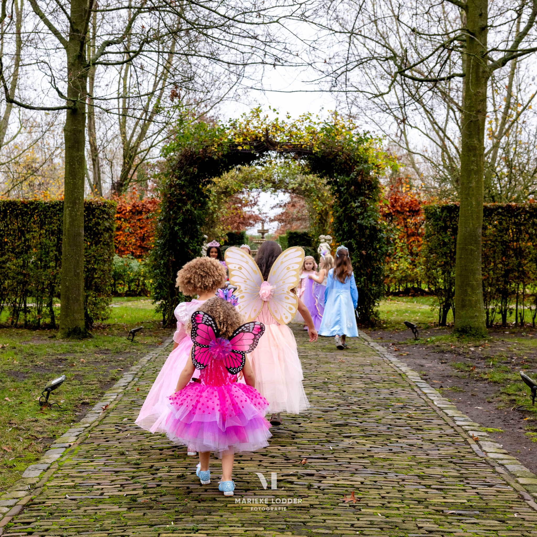 Meisje van achteren in een roze elfjesjurk met vlinderversiering, tussen andere verklede kinderen in een sfeervolle tuin.