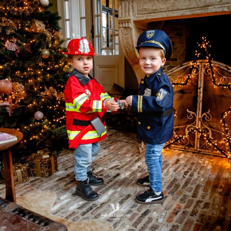 Twee jongens dragen een brandweerkostuum en donkerblauw politiepak bij een versierde kerstboom.