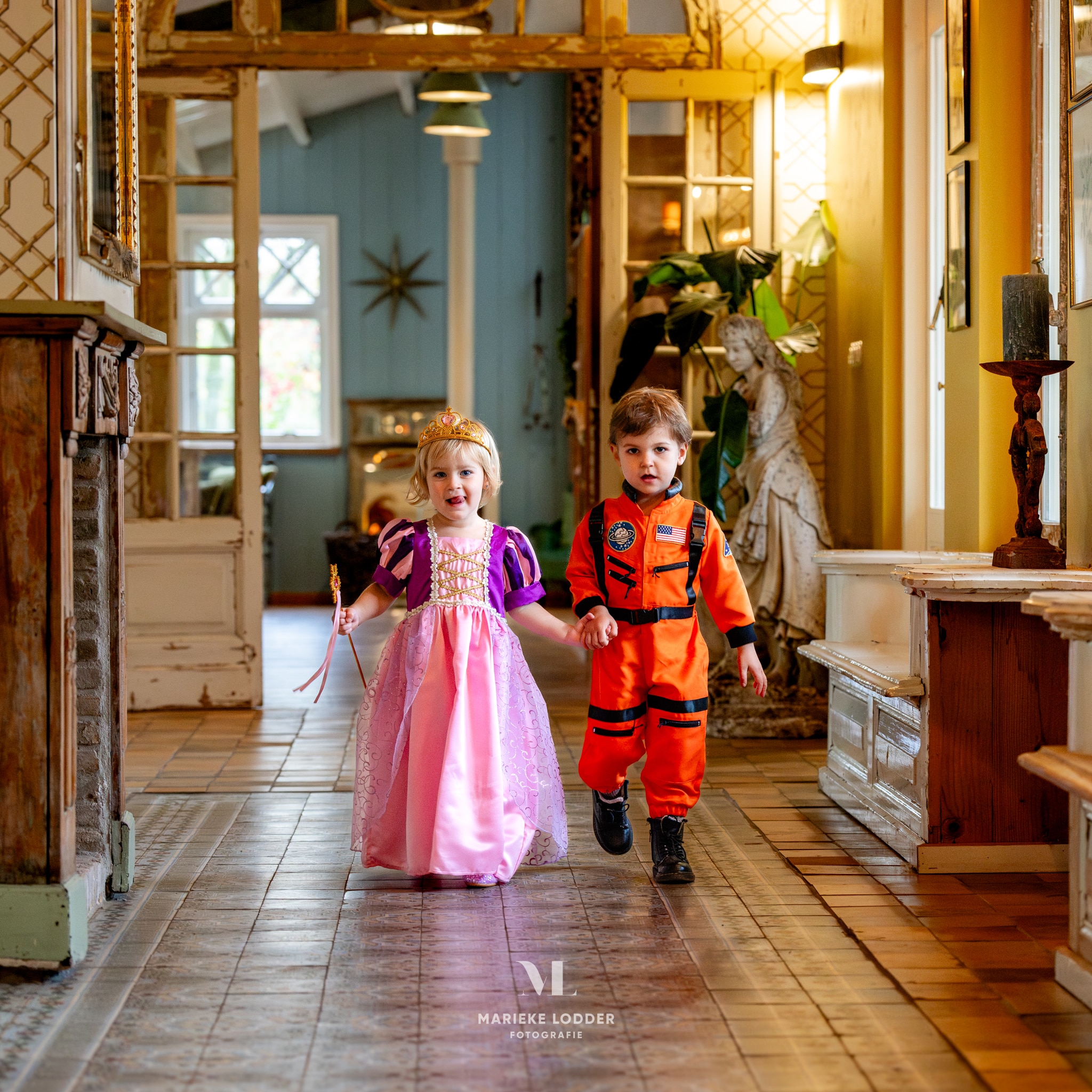 Meisje draagt een roze-paarse Rapunzel jurk met gouden details en tiara, terwijl ze hand in hand met een jongen loopt.