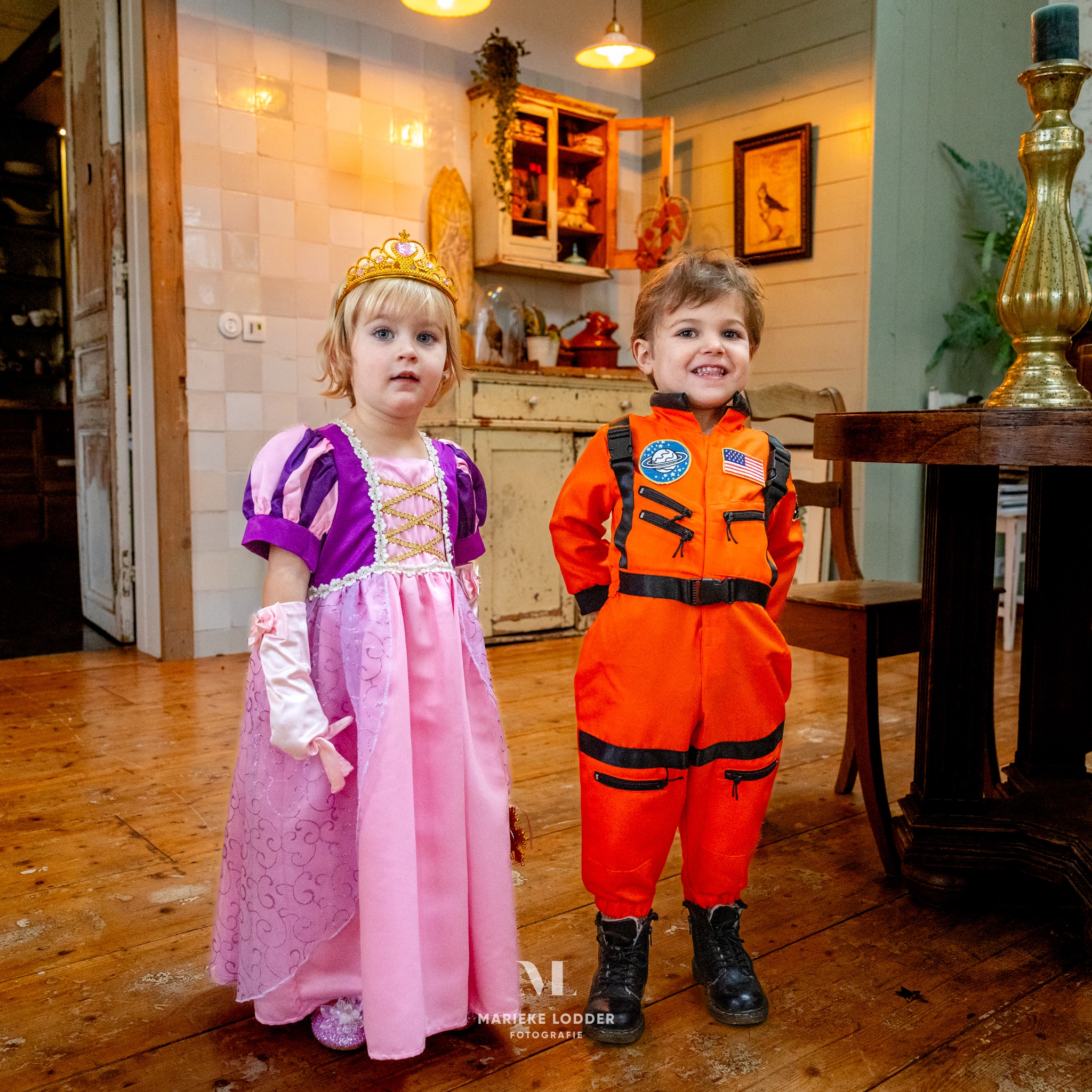 Meisje draagt de roze-paarse prinsessenjurk Rapunzel met kroon en witte handschoenen naast een jongen in astronautenpak.