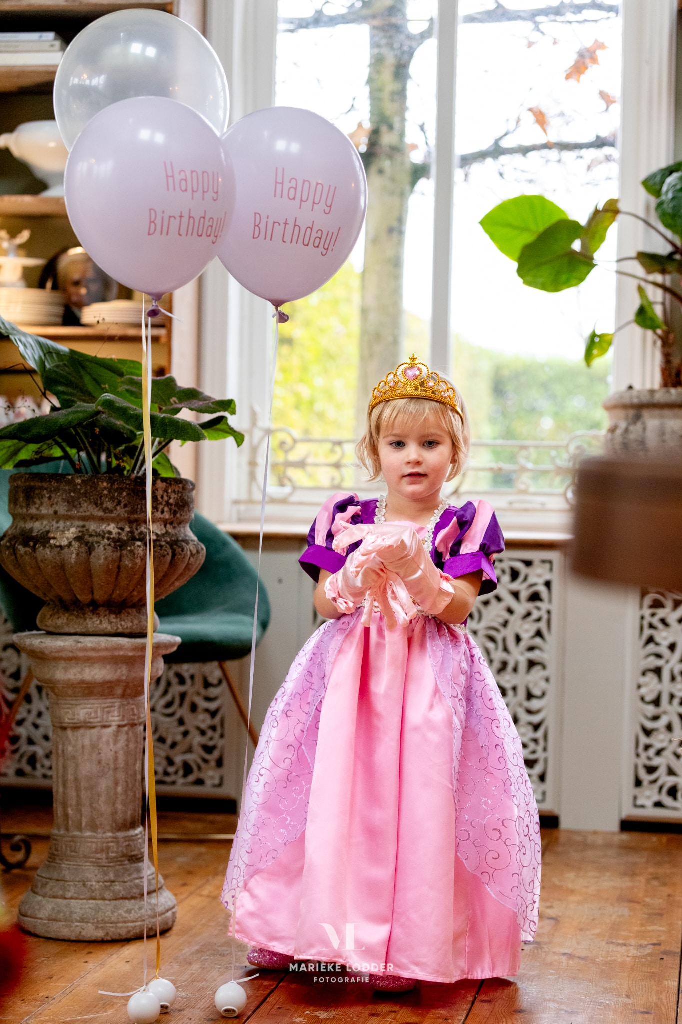 Meisje draagt een roze en paarse Rapunzel deluxe jurk met kroon en roze handschoenen in een woonkamer.