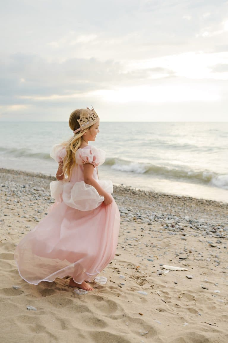 Meisje draagt een roze luxe prinsessenjurk met pofmouwen en sleep op het strand, met een gouden kroontje op haar hoofd