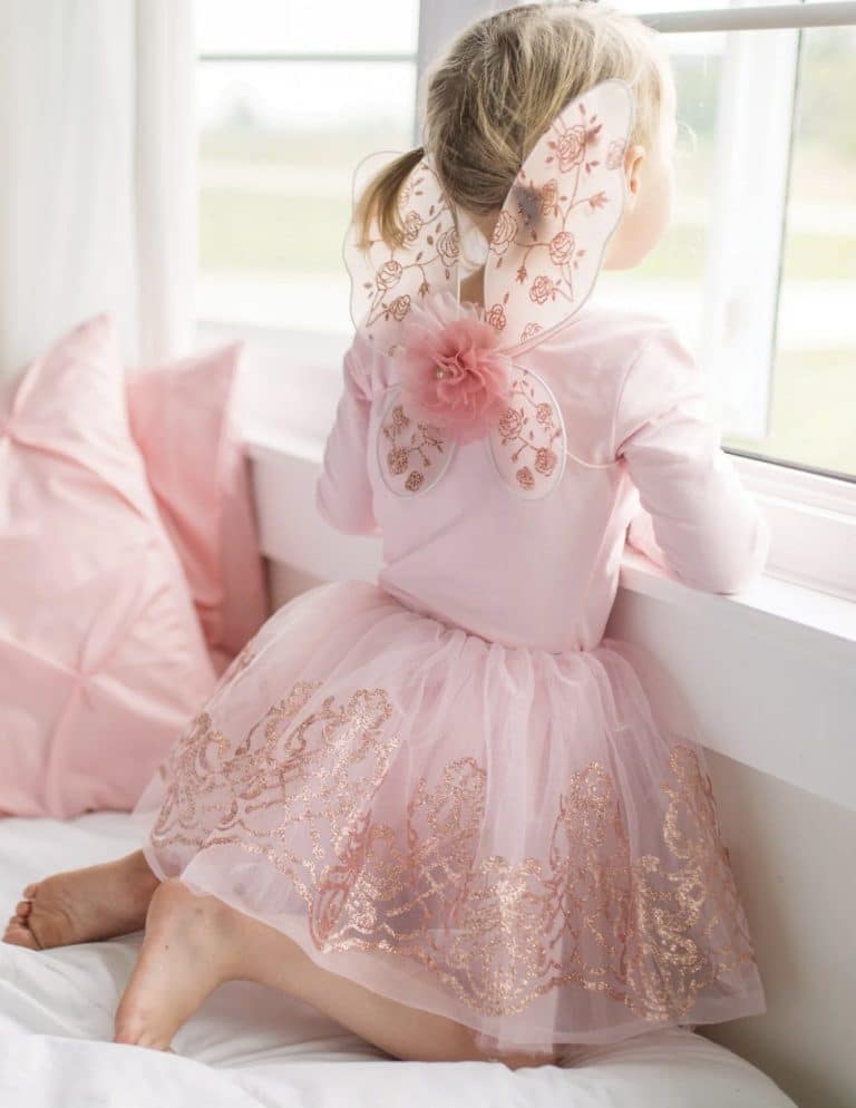 Van achteren is een kind te zien in een roze tutu rokje kind met roségouden glitterrok en sierlijke vleugels op bed bij het raam.