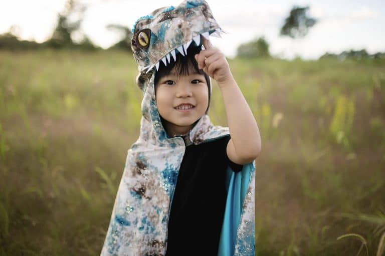 Kind draagt de blauwgroene en bruine Great Pretenders Raptor cape met dinosauruskop en puntige tanden