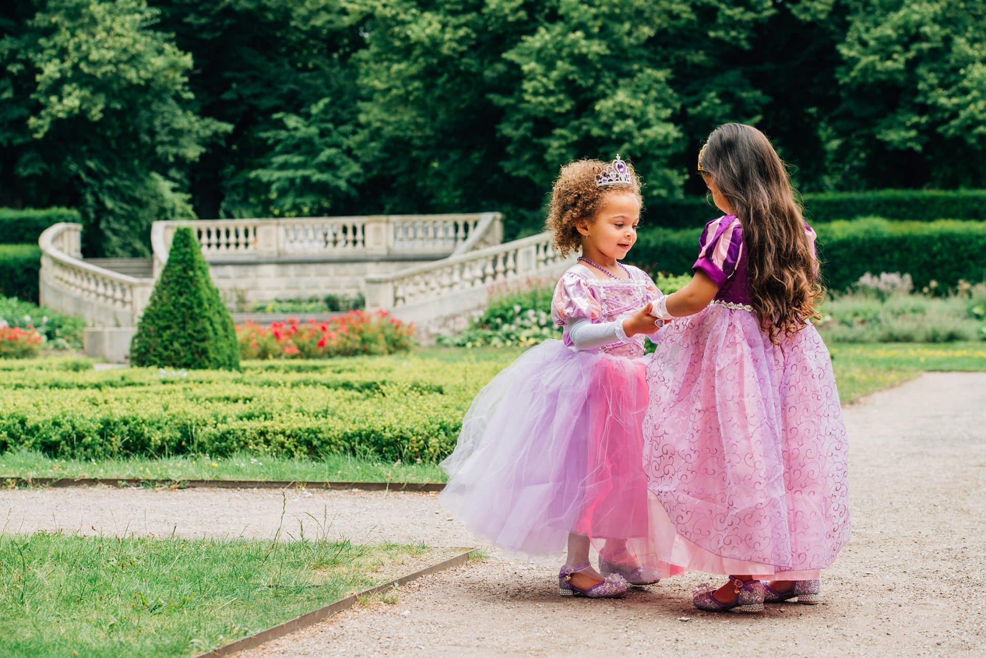 Twee meisjes dragen een roze-paarse Rapunzel prinsessenjurk in een tuin; één meisje draagt een tiara.